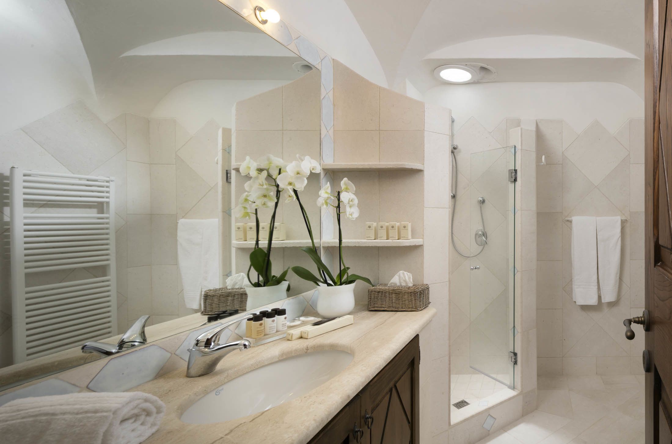A bathroom with a sink, mirror, white towels, a shower, and orchids.