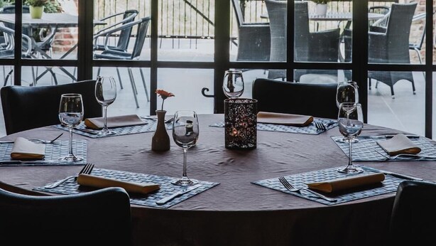 An outdoor dining area with a wooden table set for two, large windows, and a view of a cornfield. - Sparhof (Heuvelland)