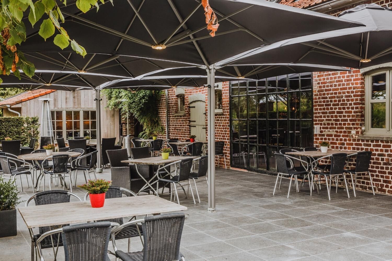Een terras buiten met tafels en stoelen, overdekt met grote parasols, en een bakstenen gebouw op de achtergrond.