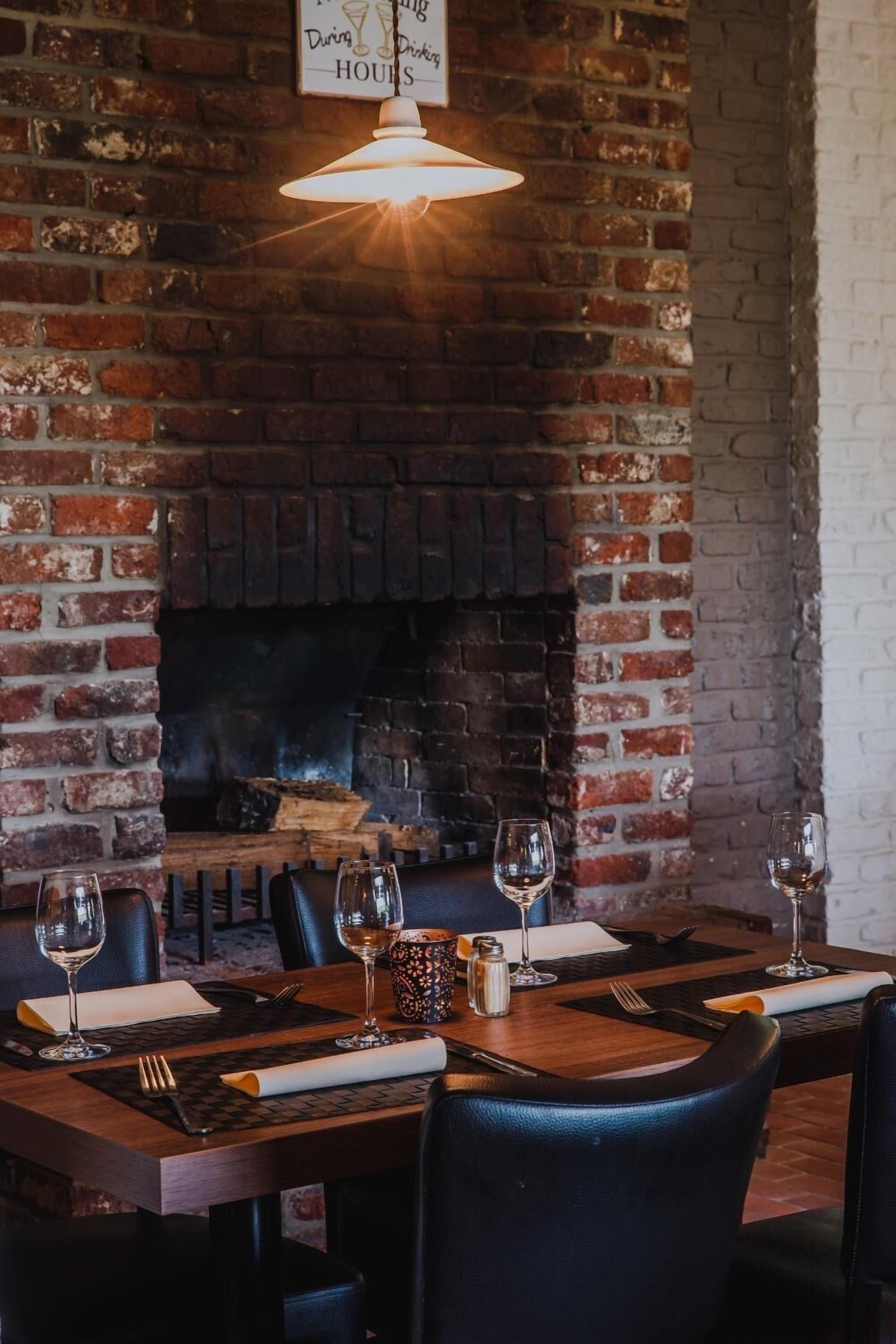 A restaurant with a brick fireplace, wooden tables, and wine glasses set for dining.