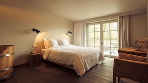 A bedroom with a bed, bedside lamps, a wooden barrel, a wicker chair, and a view through a large window. - Sparhof (Heuvelland)