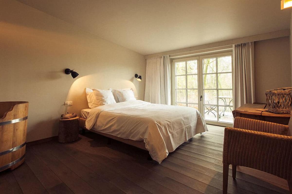 A bedroom with a bed, bedside lamps, a wooden barrel, a wicker chair, and a view through a large window.