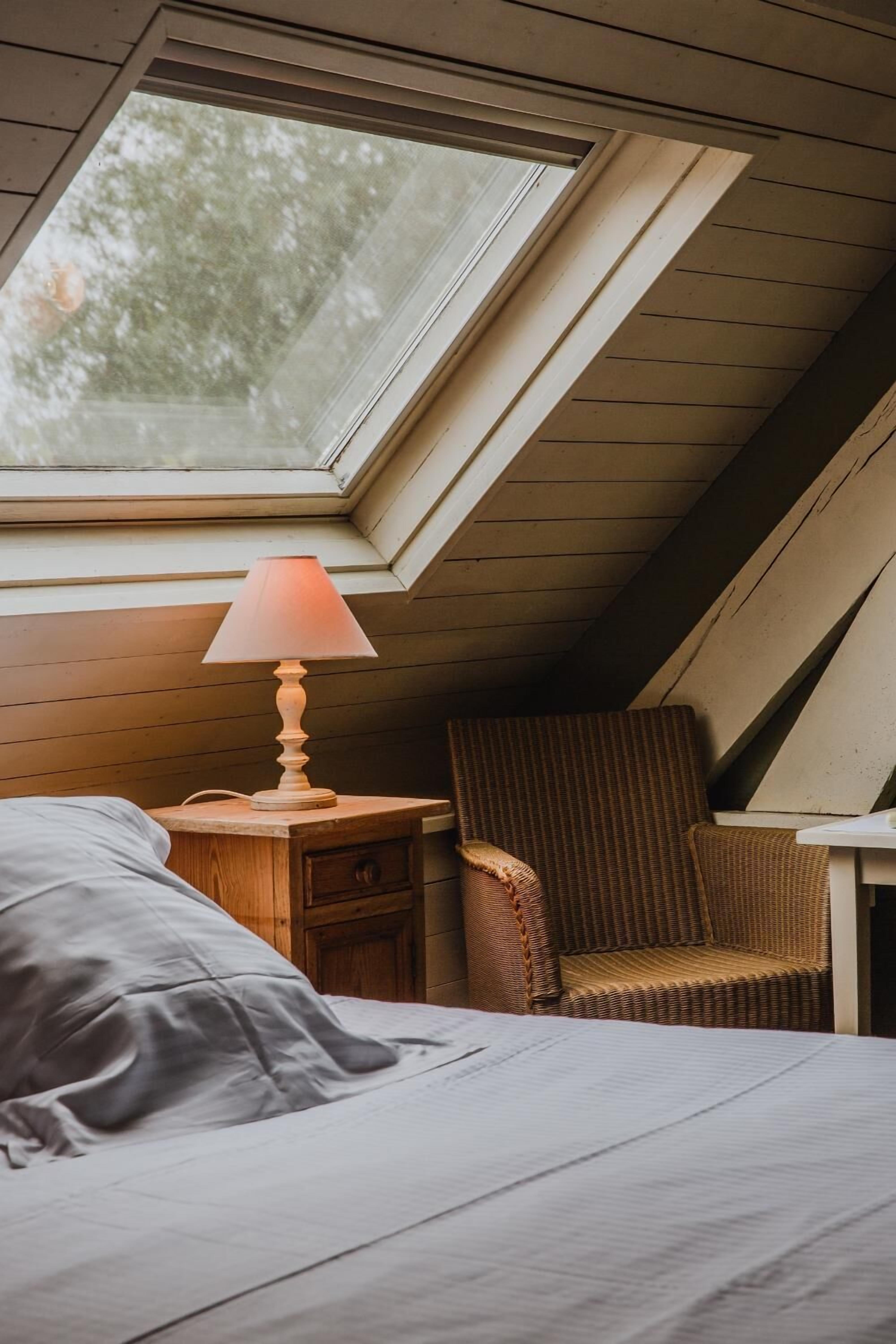 Een slaapkamer met dakraam, nachtkastje met lamp, rieten stoel en bed met grijze deken.