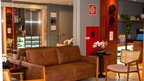 A modern hotel lobby with a brown leather sofa, wooden chairs, a small table with a vase of flowers, and a reception desk. - Nobile Hotel Juiz de Fora (Juiz de Fora)