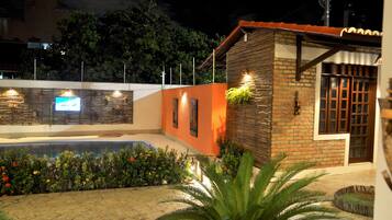 Un área de piscina con un edificio de ladrillo, un pequeño jardín con plantas y una pantalla de televisión.