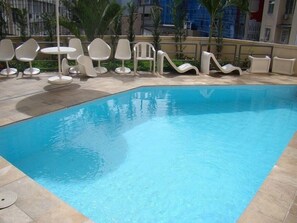 Una piscina con sedie e tavoli bianchi, circondata da un solarium piastrellato e palme.