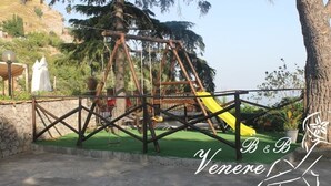 A playground with a yellow slide and a swing set, surrounded by greenery and a stone wall. - Venere B&B Castelmola (Castelmola)