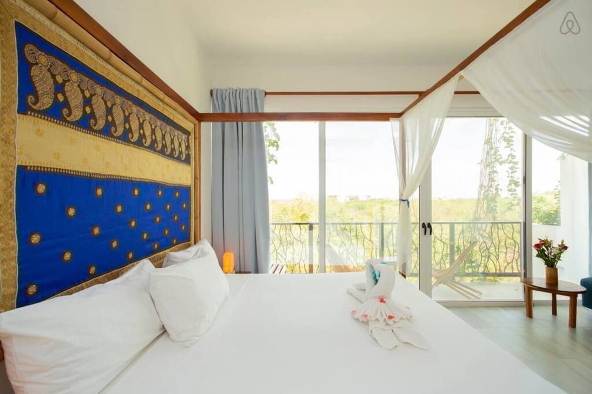 Un dormitorio amplio con una cama grande, un cabecero decorativo, un balcón con vistas y una pequeña mesa con un florero.