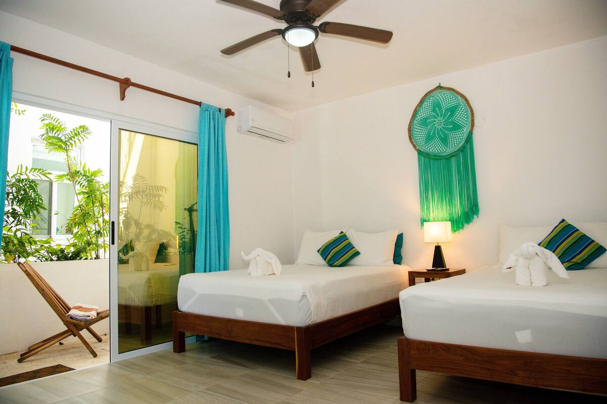 Habitación de hotel con dos camas, ventilador de techo, unidad de aire acondicionado y vista a zonas verdes a través de una puerta corrediza.
