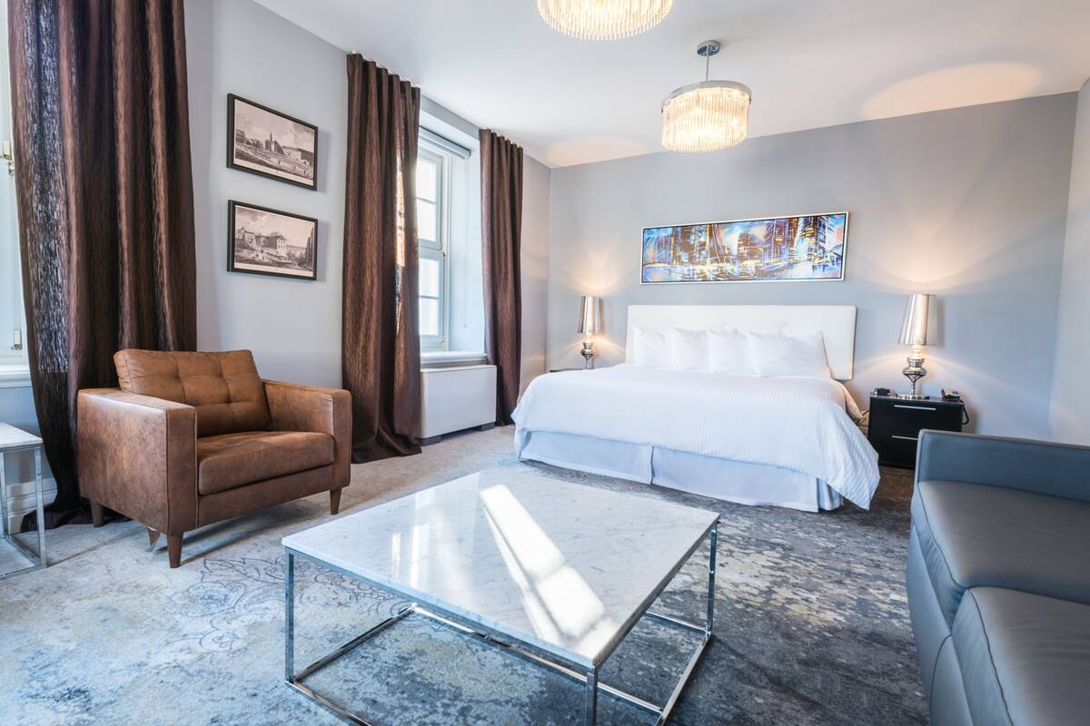 Una habitación de hotel moderna con una cama grande, una mesita de centro de vidrio, un sillón de cuero marrón y un sofá gris.