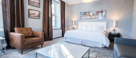 A modern hotel room with a large bed, a glass coffee table, a brown leather armchair, and a grey sofa.