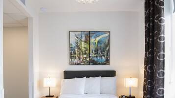 A hotel room with a large bed, two bedside lamps, a chandelier, a painting on the wall, and a window with curtains.