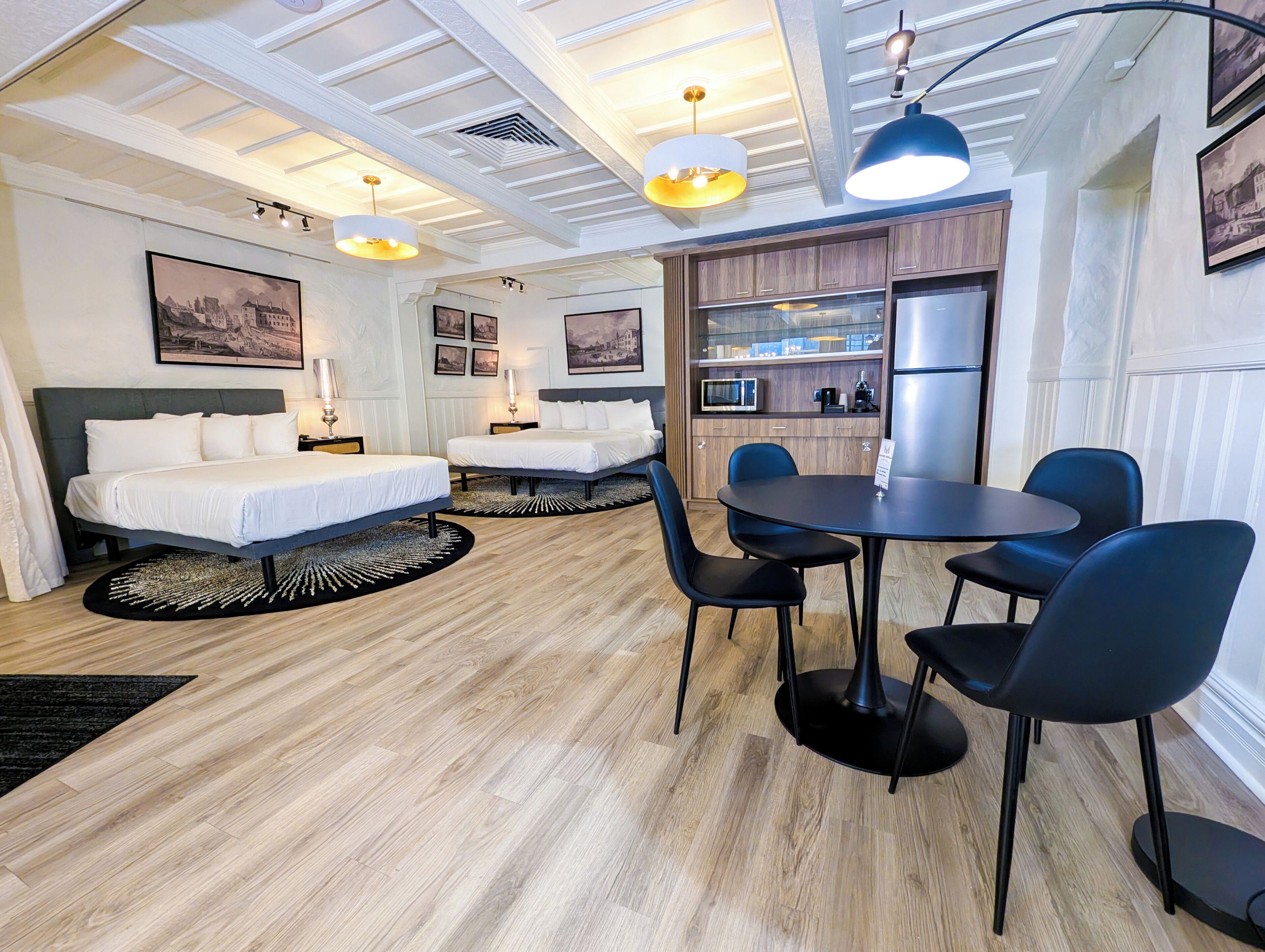 A hotel room with two beds, a dining table with chairs, a kitchen area, and a ceiling with recessed lighting.