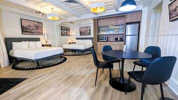 A hotel room with two beds, a dining table with chairs, a kitchen area, and a ceiling with recessed lighting.