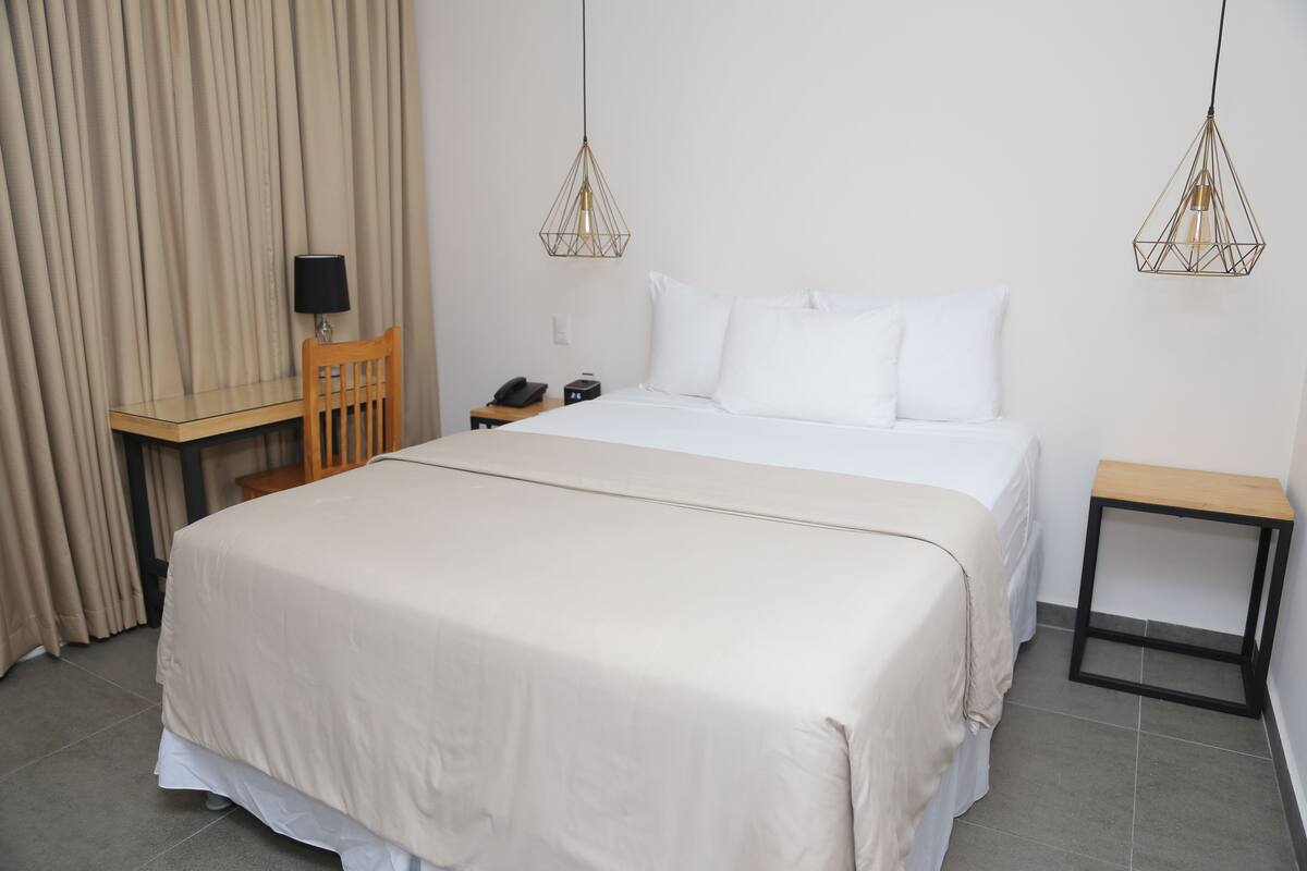 A neatly arranged hotel room with a large bed, a desk, a chair, and two pendant lights.