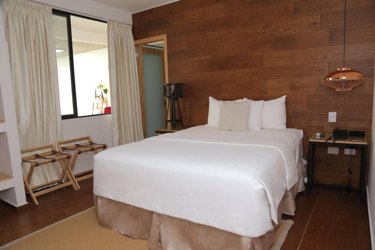 A bedroom with a large bed, wooden flooring, a wooden headboard, a bedside table with a lamp, and a window with curtains.