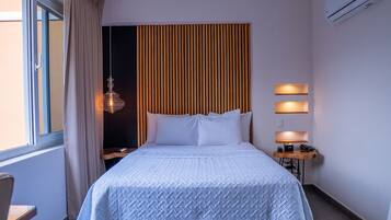 Quarto de hotel com uma cama grande, cabeceira de madeira, escrivaninha com abajur e ar condicionado.
