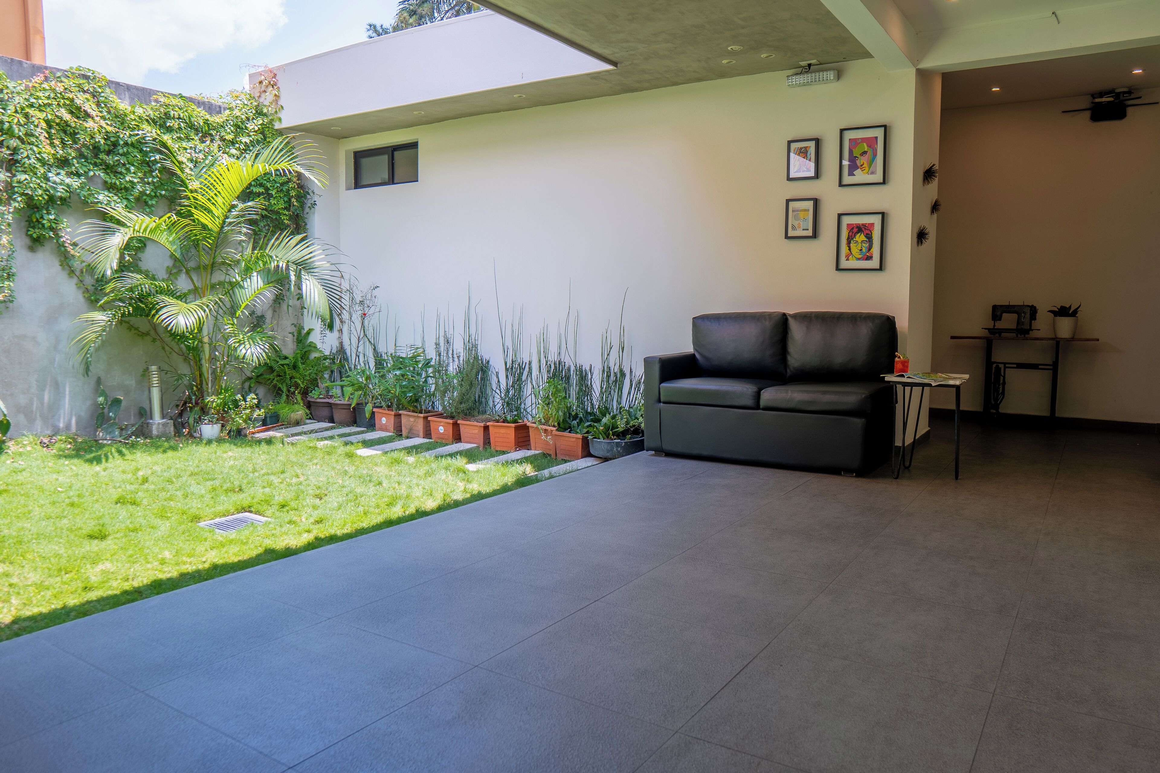 Zona d'estar moderna amb un sofà gris, una taula petita i un televisor fixat a la paret. Hi ha un jardí amb diverses plantes i un camí pavimentat.