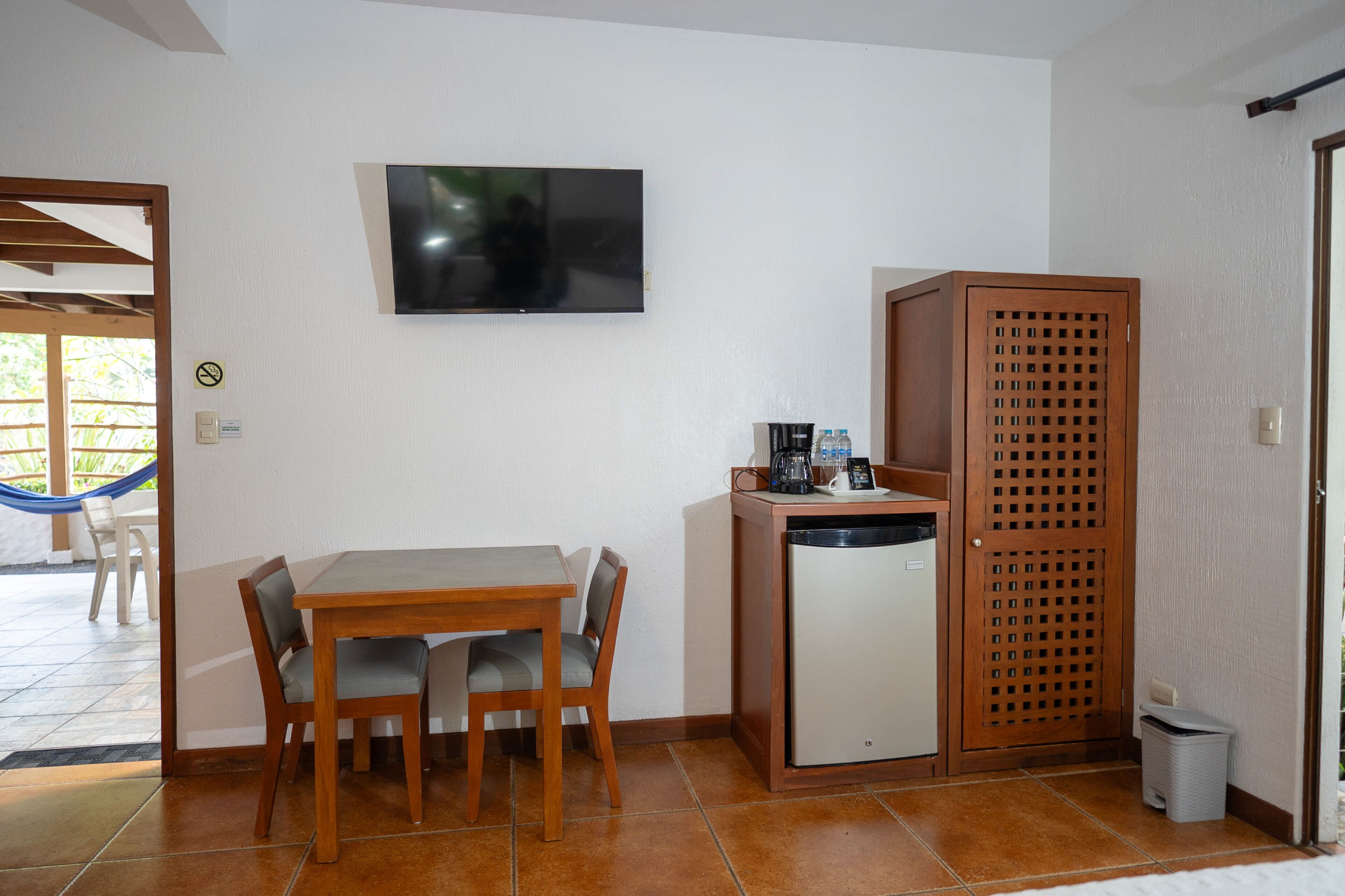 Una habitación compacta con una mesa y sillas de madera, un refrigerador pequeño, un televisor montado en la pared y un mueble de madera con diseño de celosía.