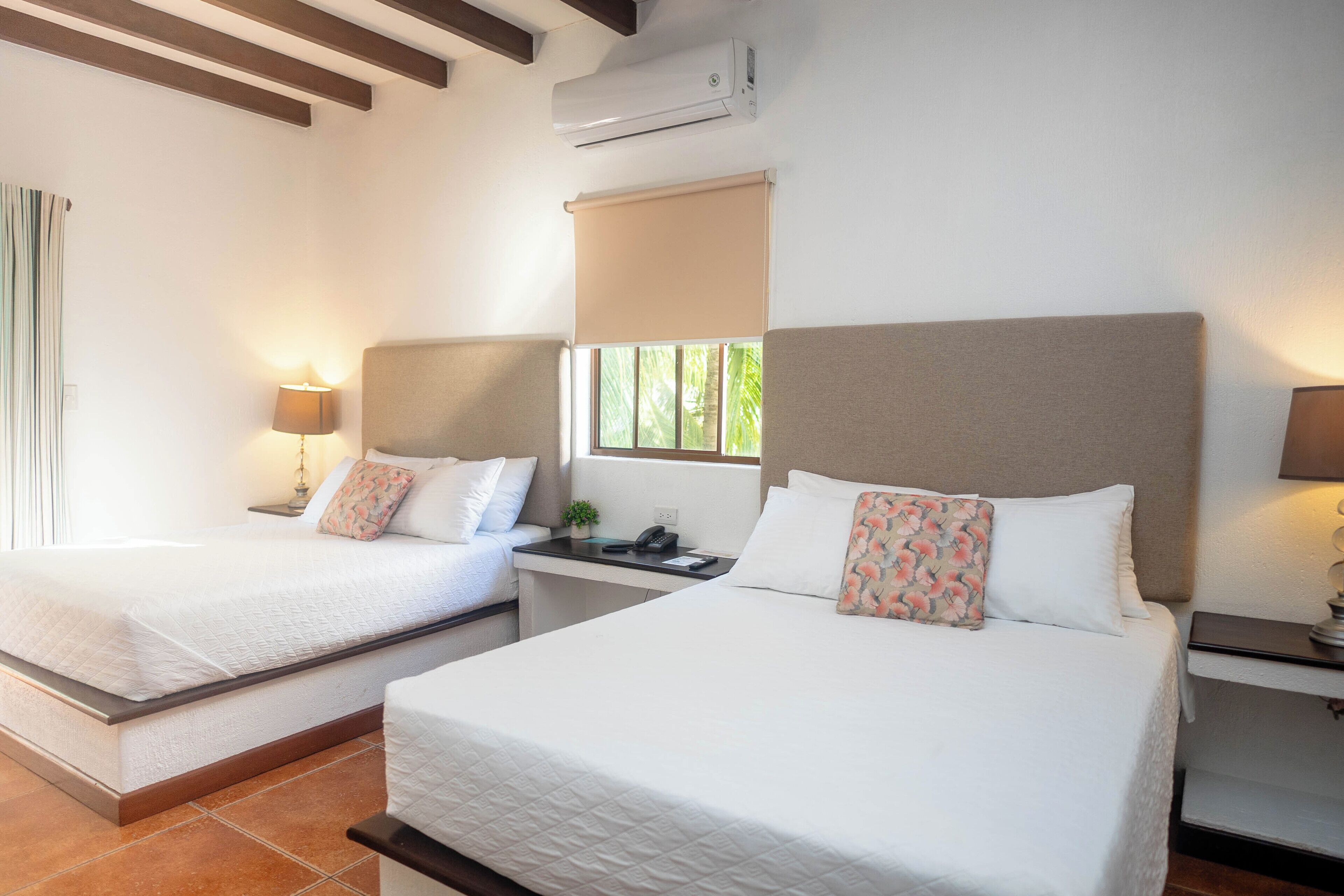 A hotel room with two beds, a desk, and a window with a view of greenery.