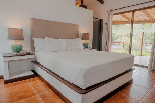 A neatly made bed with white linens, two bedside lamps, and a view of the outdoors through a sliding glass door.