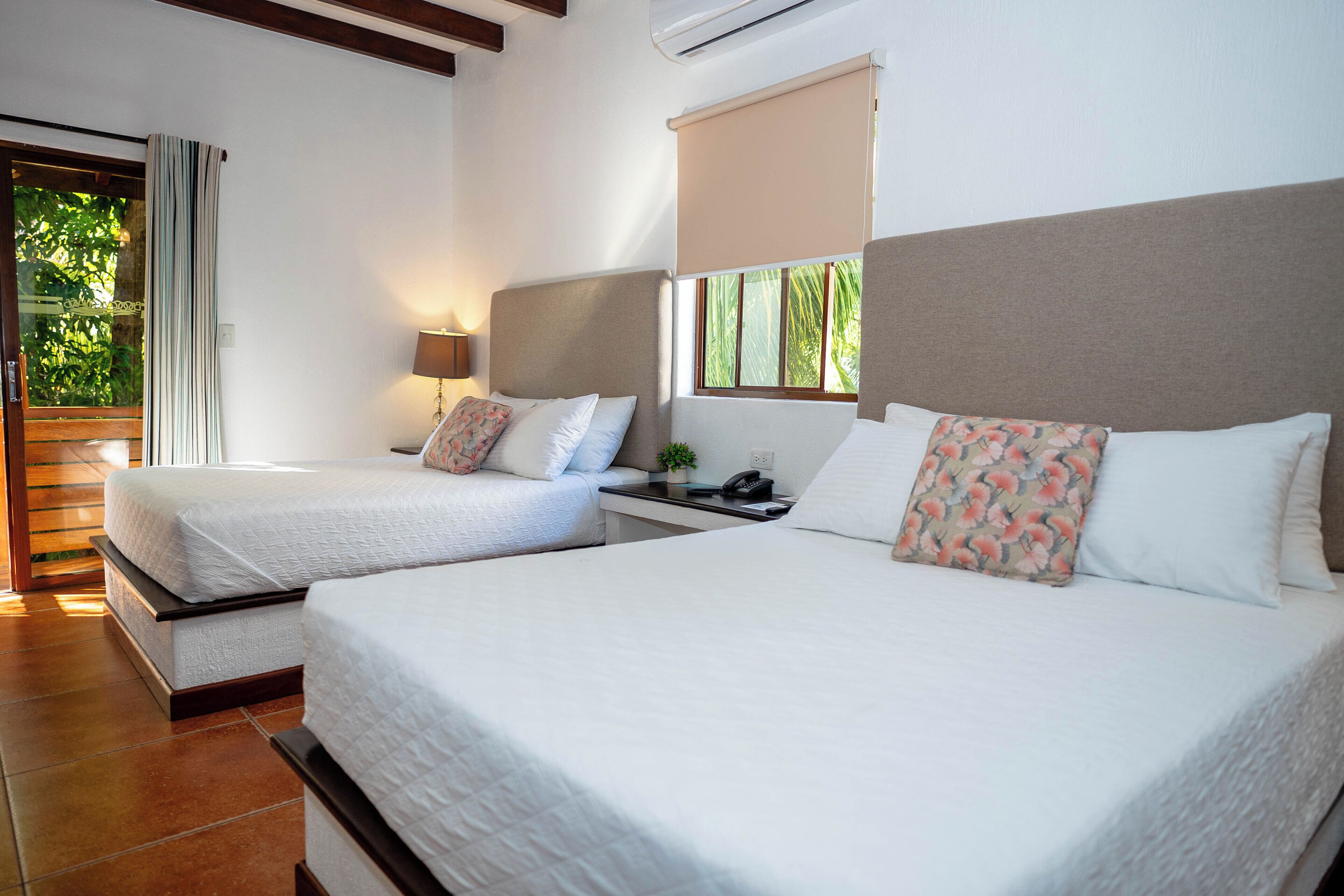 Habitación de hotel con dos camas, una mesita de noche con lámpara, teléfono y ventana con vistas a zonas verdes.
