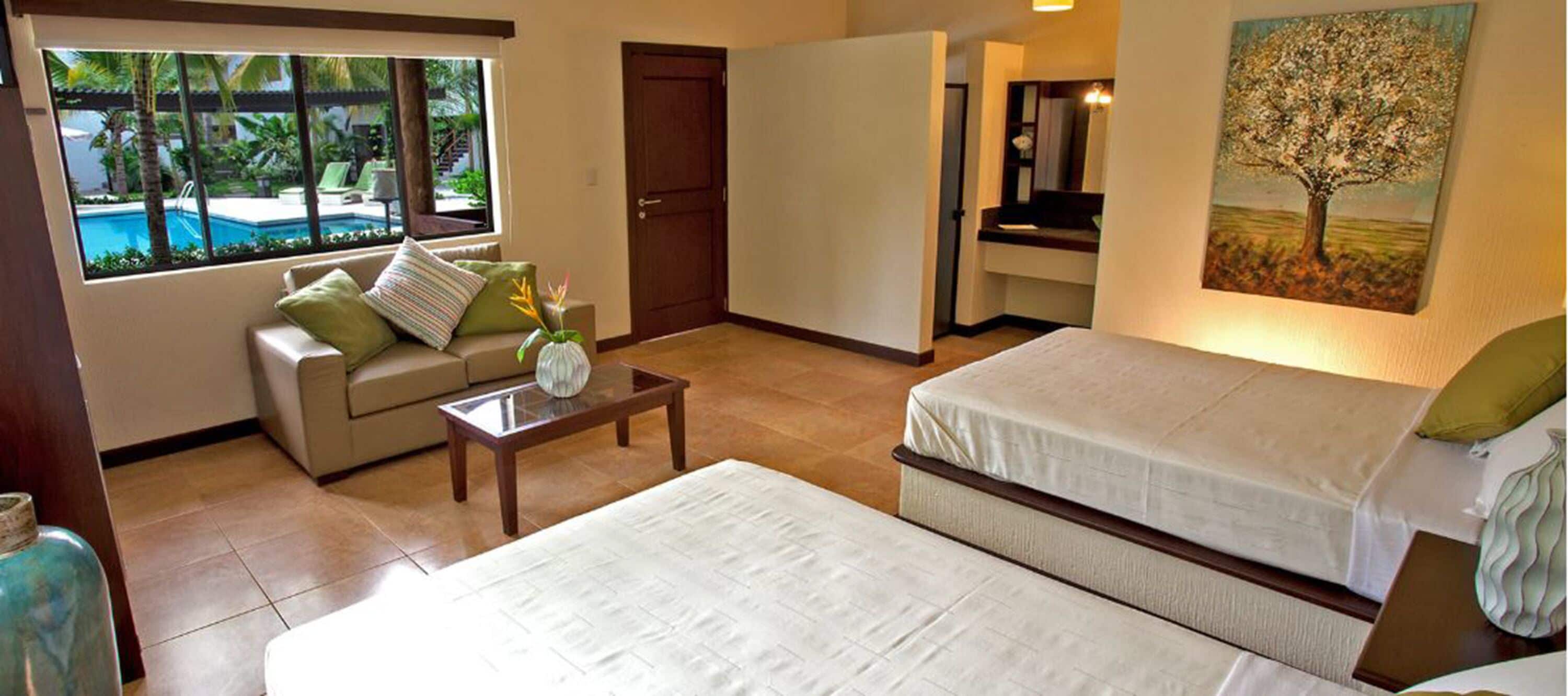 Habitación de hotel con cama, sofá, una mesita, un cuadro y vistas a una piscina y zonas verdes.