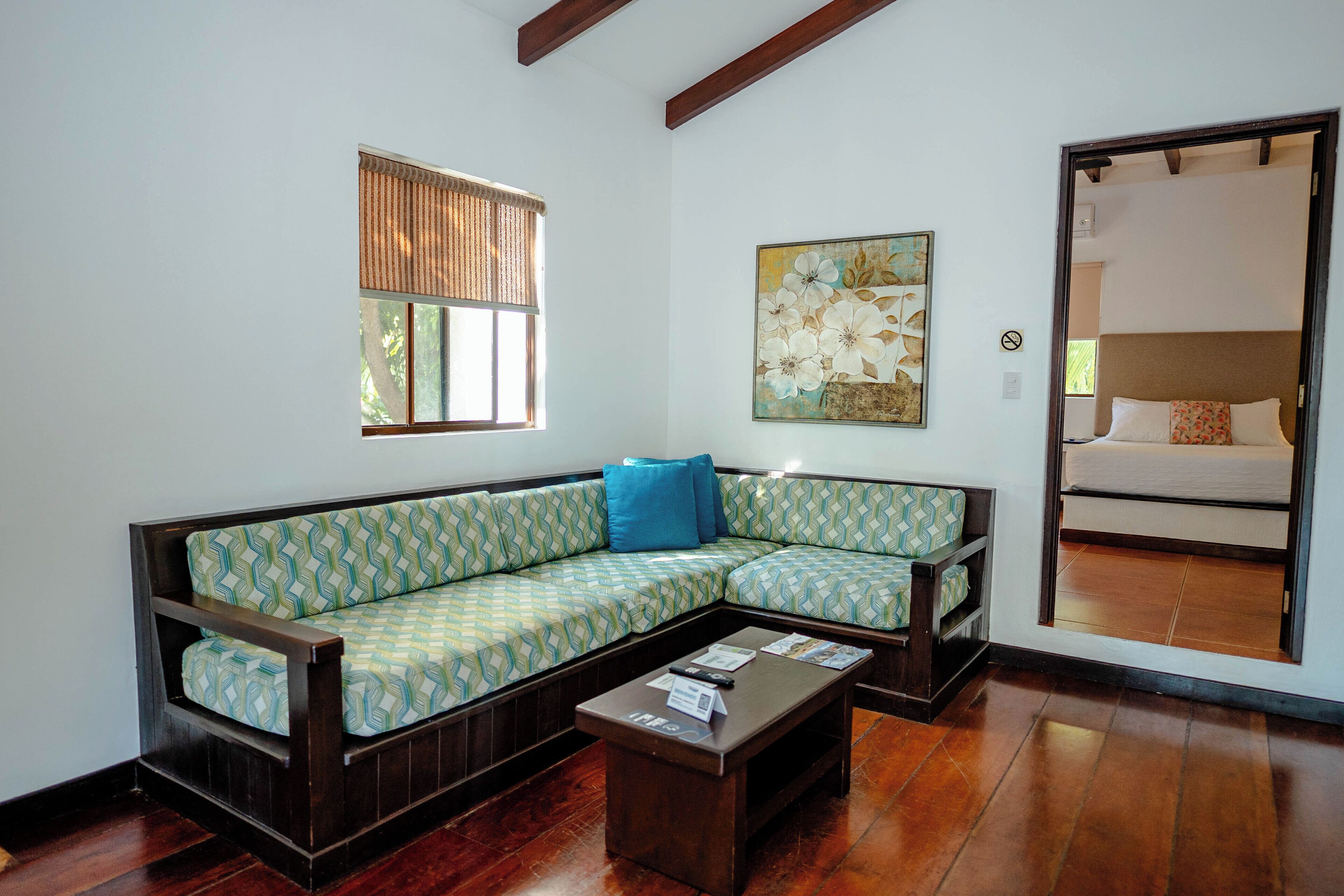 A room with a sectional sofa, a coffee table, a window with blinds, and a painting on the wall.