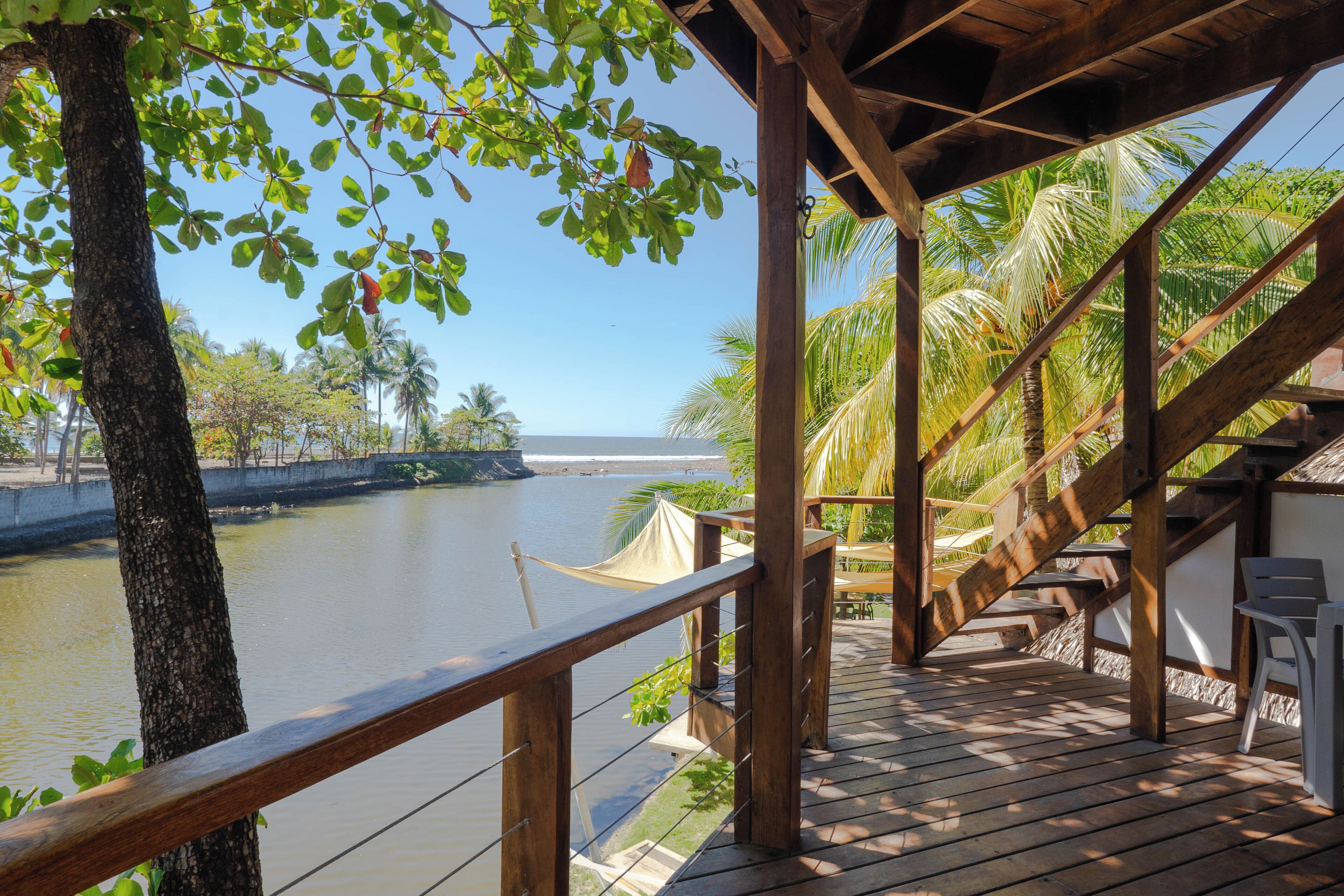 Una terraza de madera con vista a un río tranquilo, con una estructura de techo de paja y mobiliario exterior.