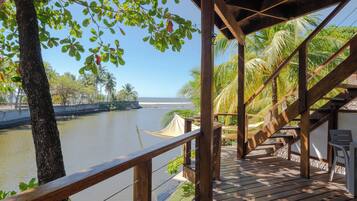 Una terraza de madera con vista a un río tranquilo, con una estructura de techo de paja y mobiliario exterior.