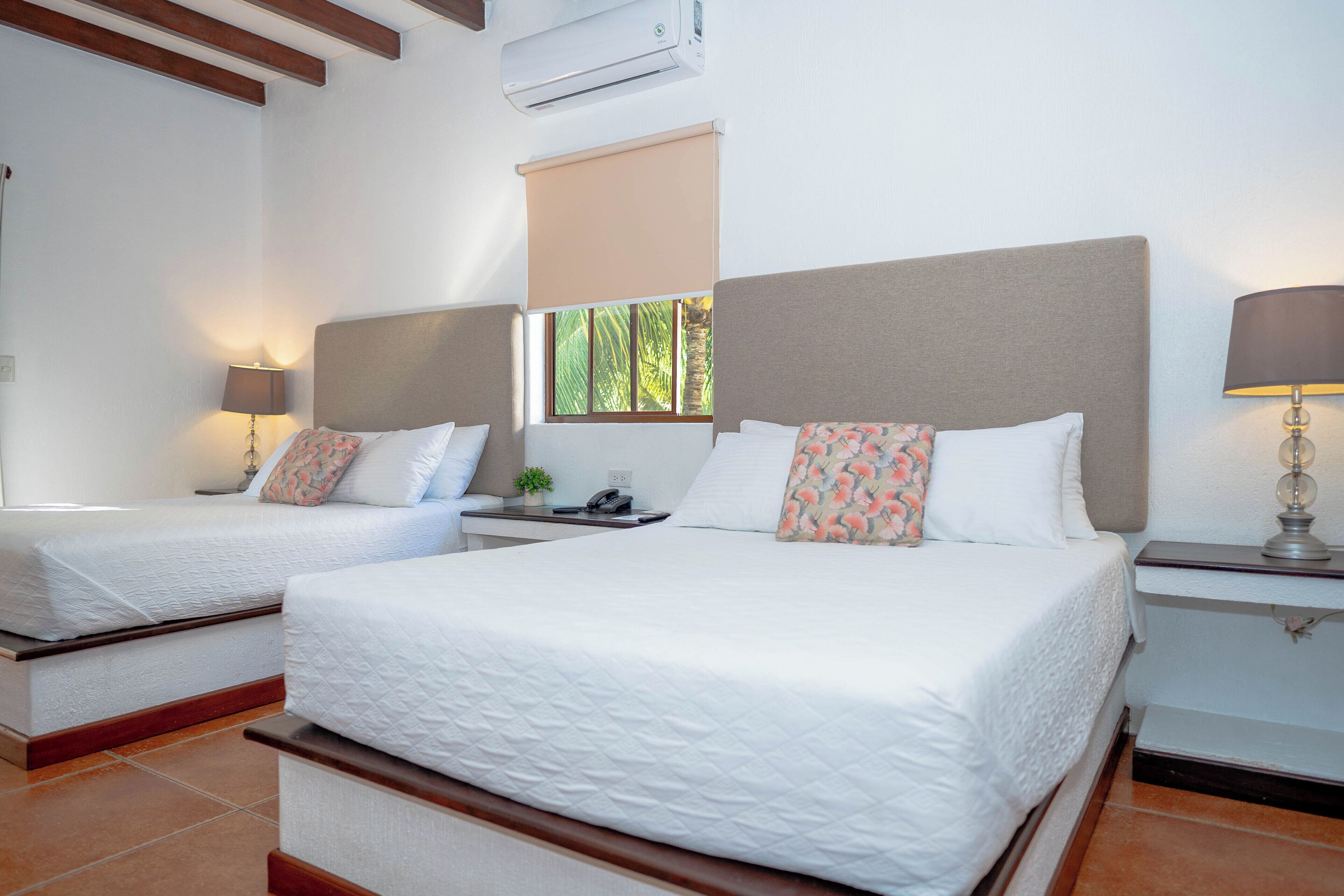 A hotel room with two beds, a window with a view of greenery, a bedside table with a lamp, and an air conditioning unit on the wall.