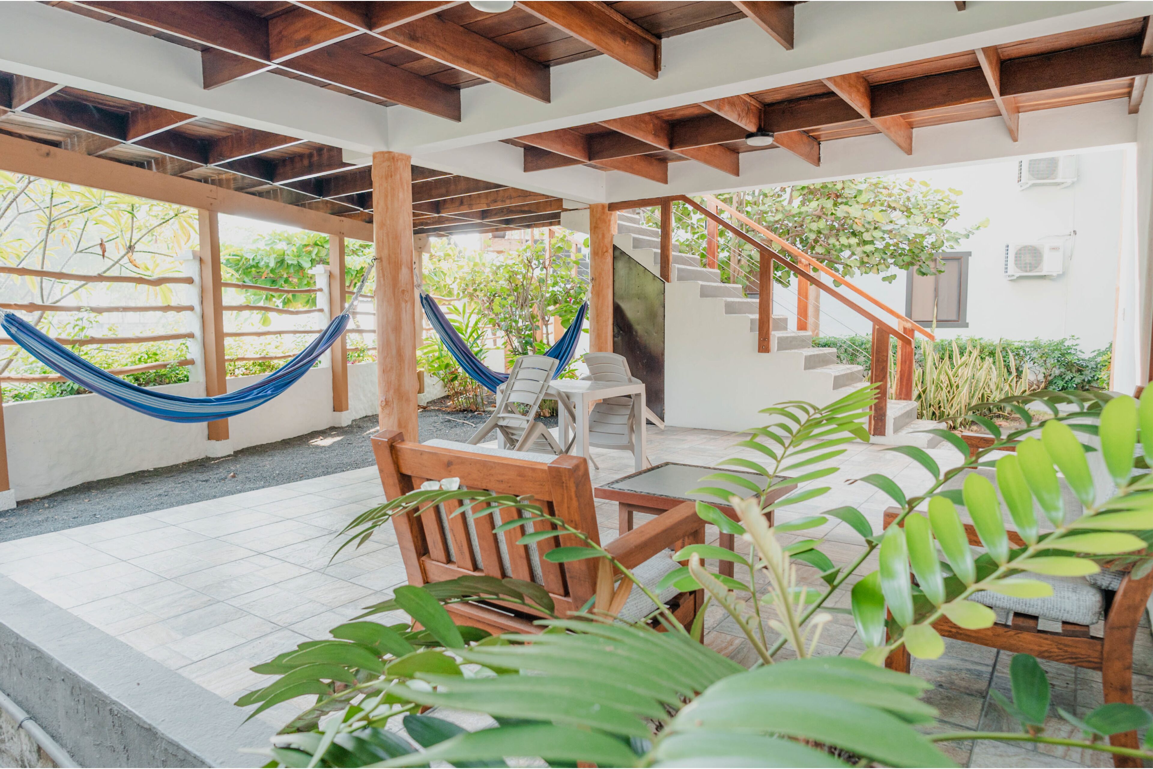 A covered patio area with wooden furniture, a hammock, and a staircase leading to an upper level.