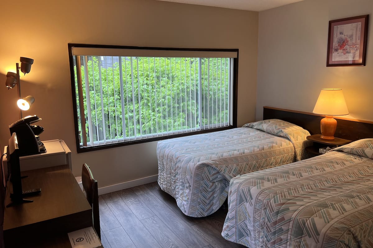 A hotel room with two beds, a desk with a lamp, a chair, and a window with blinds.