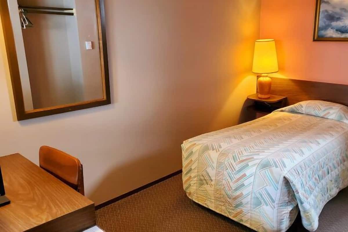 A hotel room with a bed, a desk, a chair, a nightstand with a lamp, a painting on the wall, and a mirror.