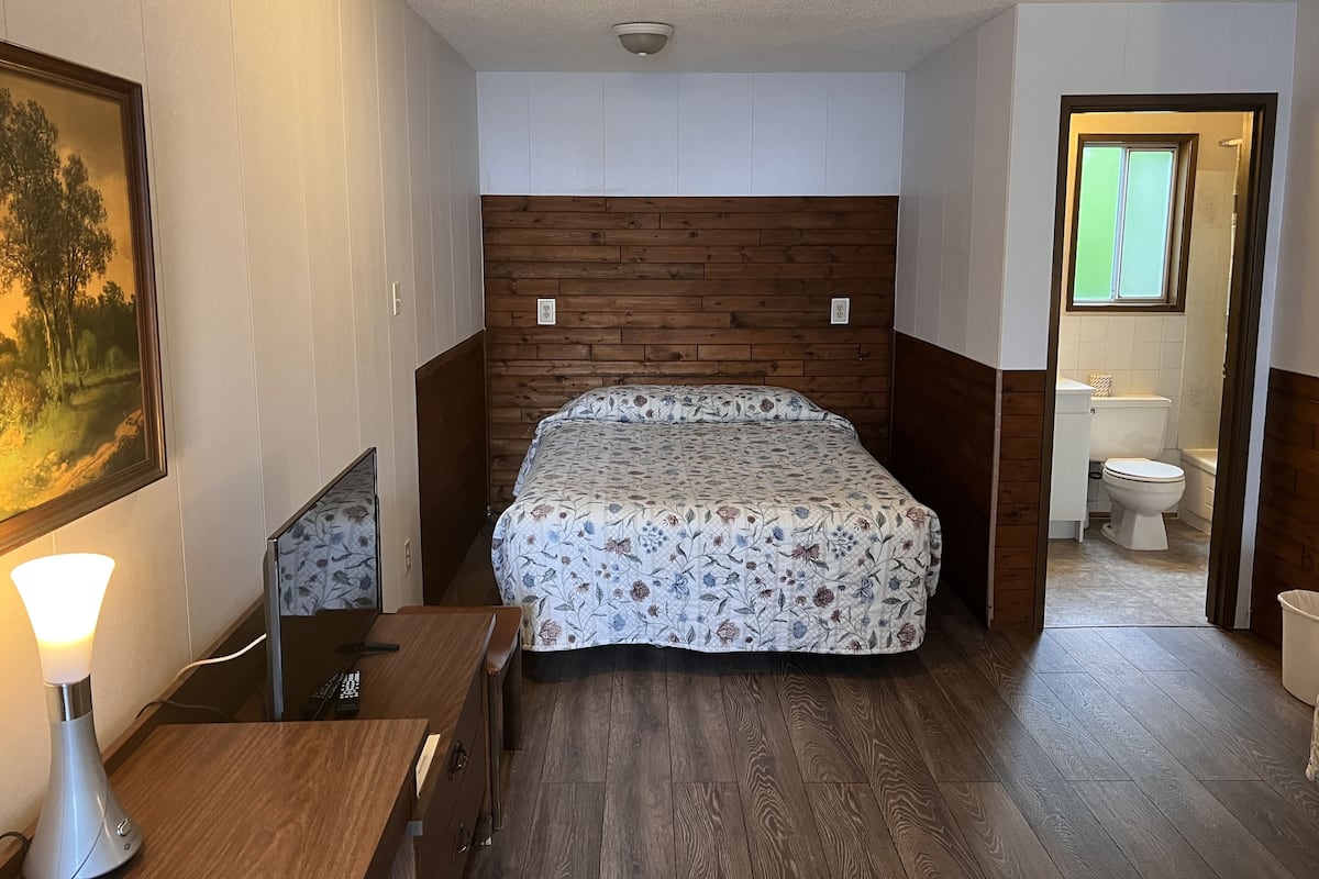 A bedroom with a wooden headboard, a bed with floral bedding, a desk with a lamp, and a view of the bathroom.