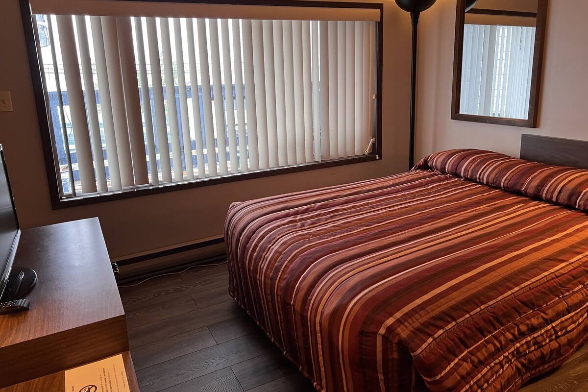 A hotel room with a large window with blinds, a bed with striped bedding, a bedside table with a lamp, and a mirror on the wall.