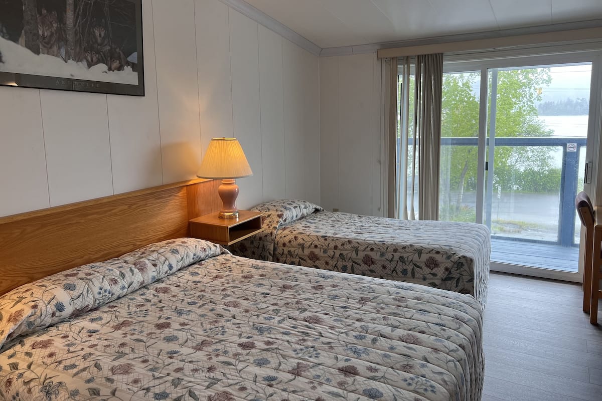 A hotel room with two beds, a nightstand, a lamp, and a large window with a view of the outdoors.