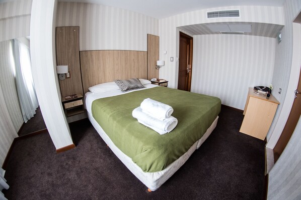 Habitación de hotel con una cama grande, cabecera de madera, mesita de noche, lámpara y un pequeño mueble de madera.