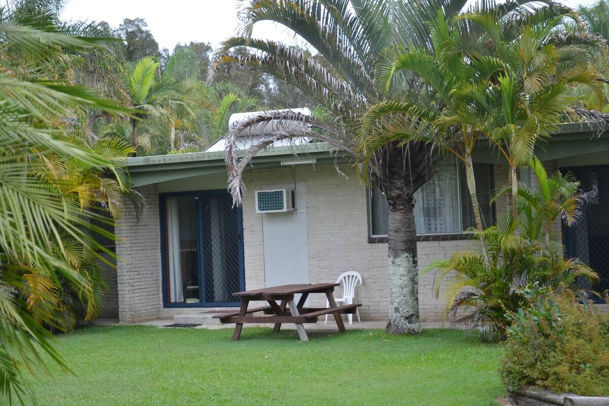 A patio with a picnic table and chairs, surrounded by palm trees and a building with a green roof and a window with a metal grid.