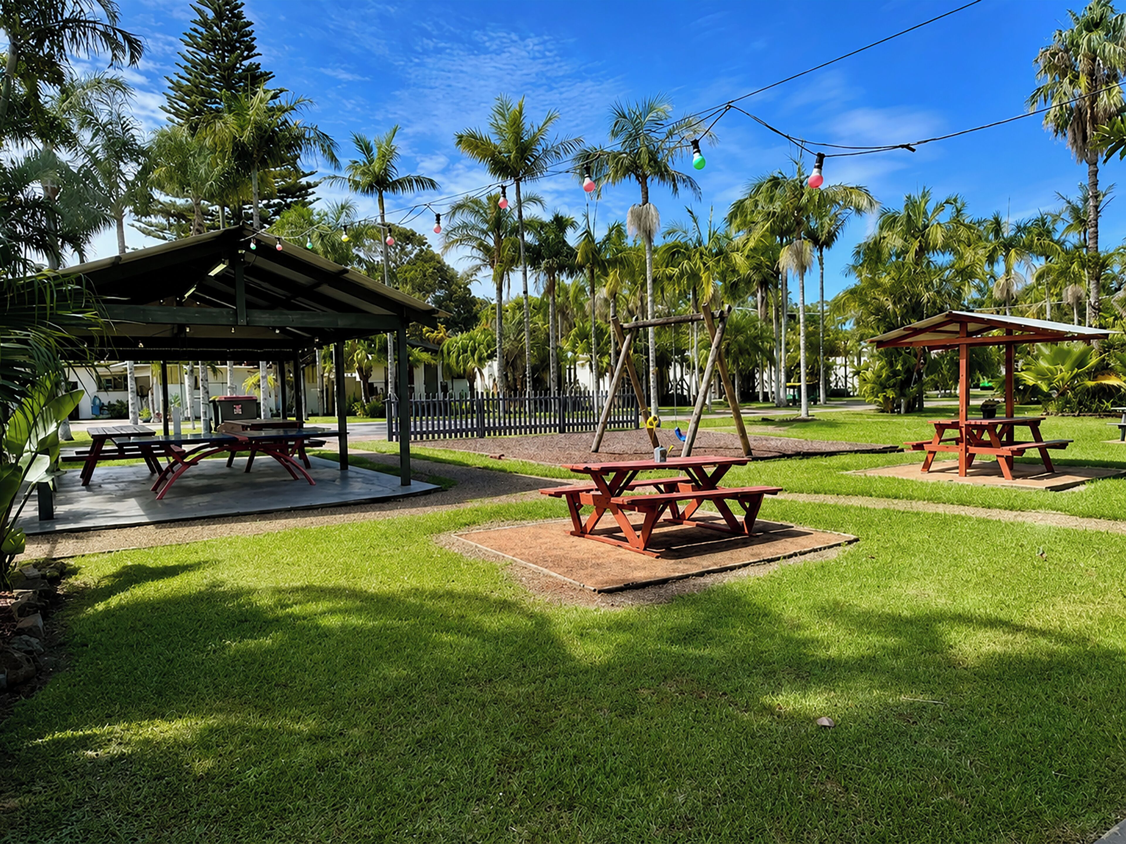 A park with picnic tables and a swing set, surrounded by palm trees and a clear sky.