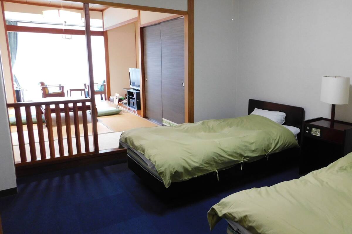 A hotel room with two beds, a desk, a chair, and a sliding door leading to a balcony.