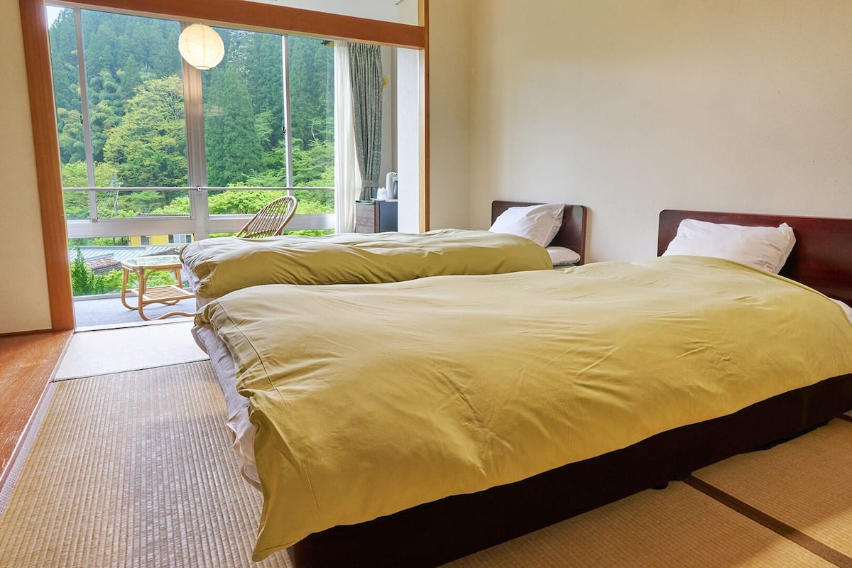 A room with two beds, a view of greenery outside, and a chair by the window.