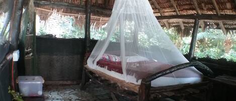 A rustic room with a bed under a mosquito net, a small table with a container, and a tiled floor.