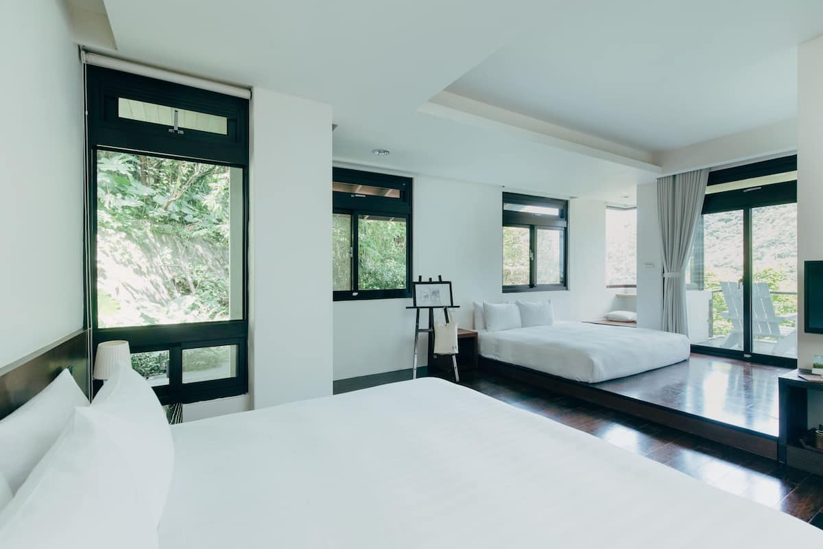 Una habitación de hotel moderna con una cama grande, una cama más pequeña y un escritorio con una silla. Amplias ventanas ofrecen vistas del exterior.