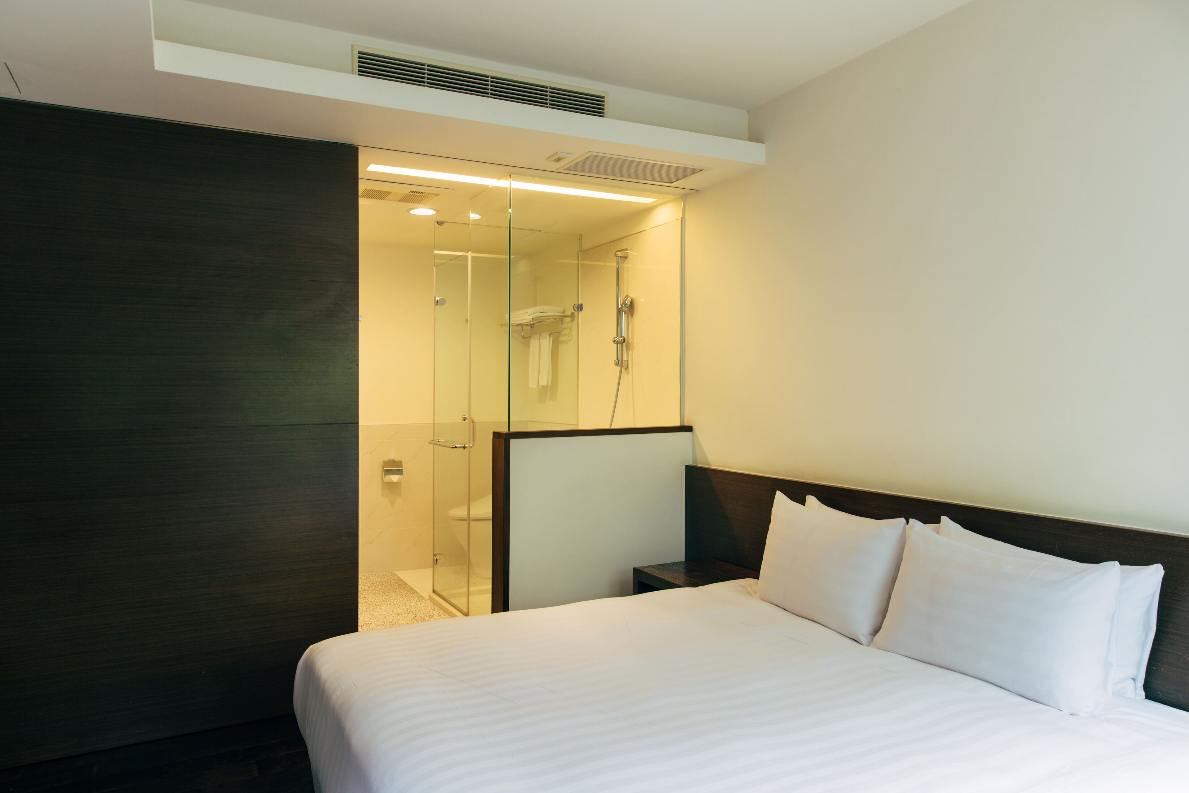 A hotel room with a large bed, a dark wooden headboard, a glass shower enclosure, and a small bedside table.