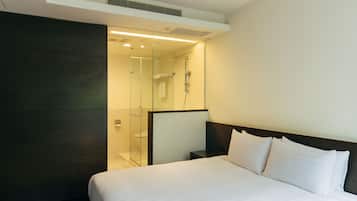 A hotel room with a large bed, a dark wooden headboard, a glass shower enclosure, and a small bedside table.