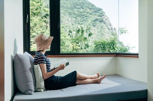 Una persona sentada en un banco junto a una ventana con vista a una montaña y follaje.