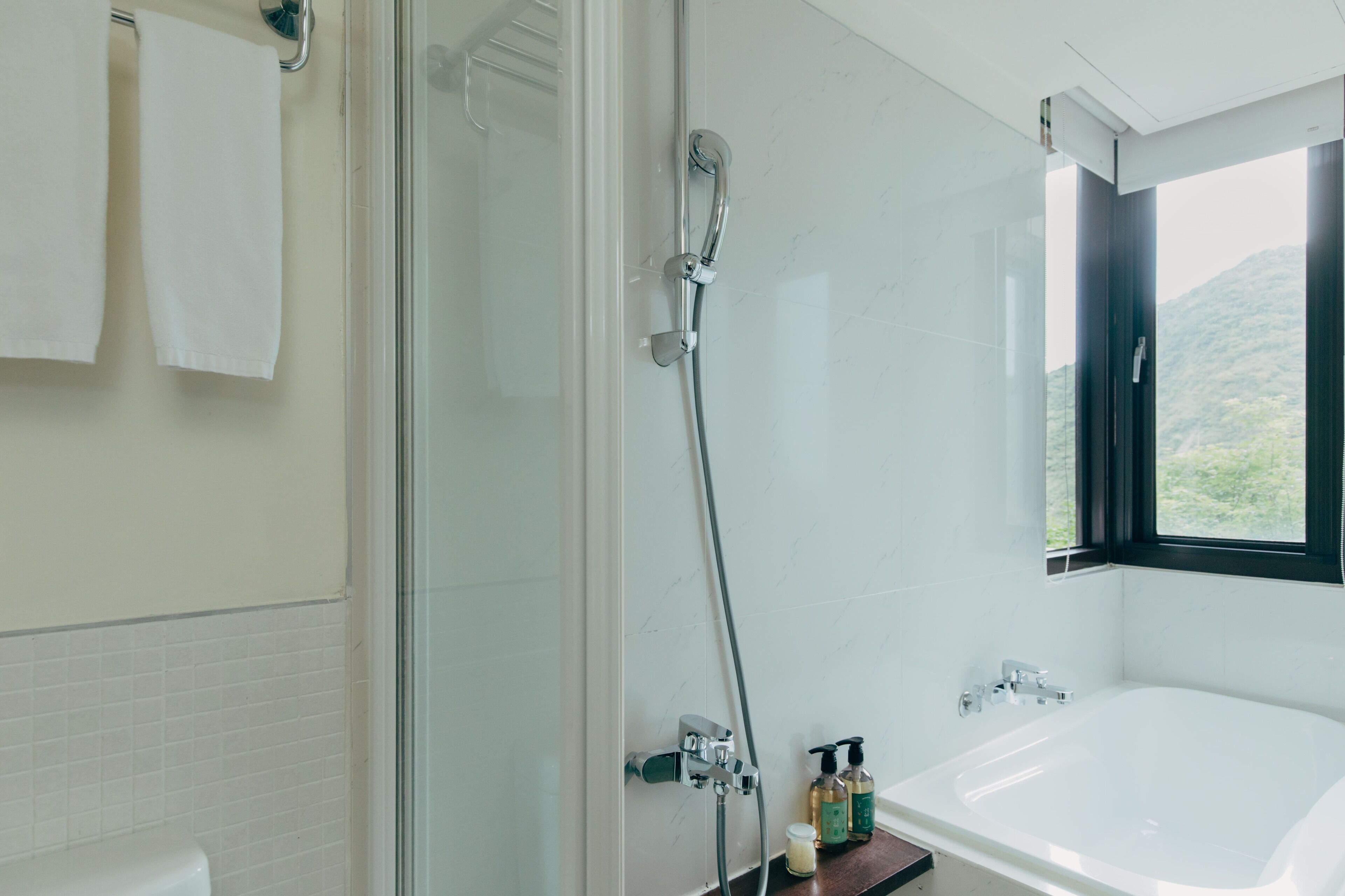 A modern bathroom with a glass-enclosed shower, a white bathtub, and a window offering a view of greenery.