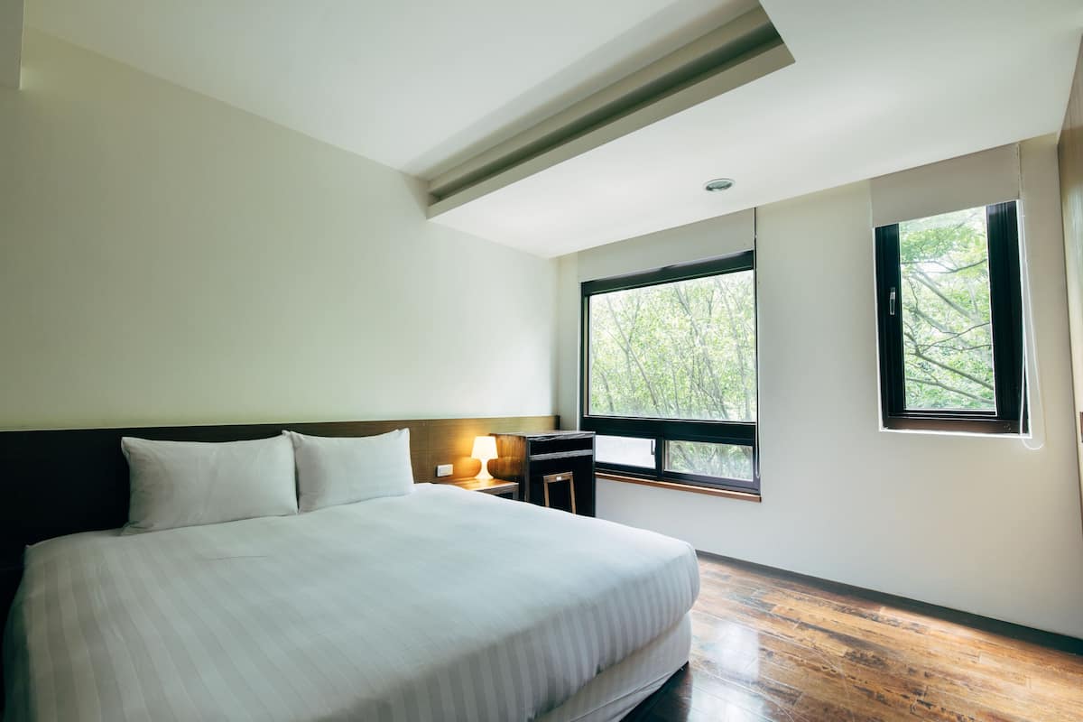 Habitación de hotel con una cama grande, una mesita de noche con una lámpara y una ventana con vistas a árboles.