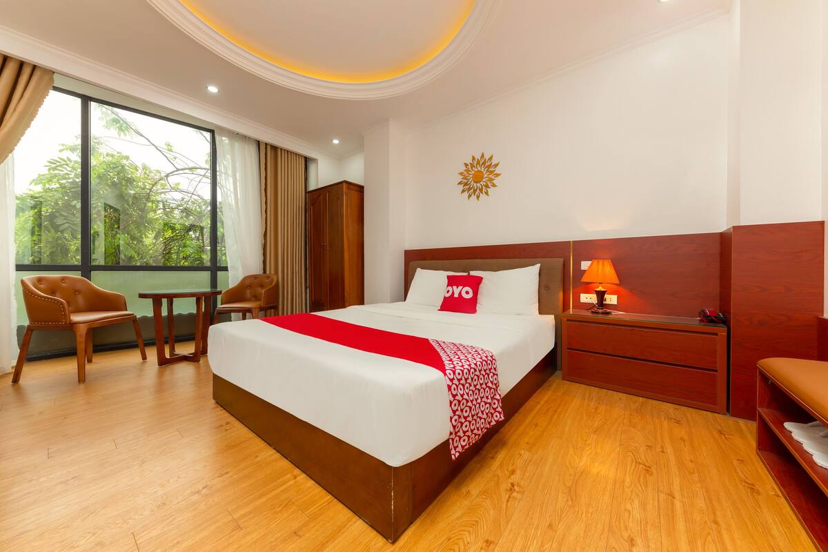 Habitación de hotel con una cama grande, muebles de madera, una mesita de noche con lámpara y vistas a zonas verdes por la ventana.